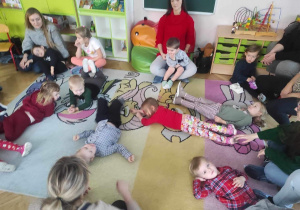 Dzieci leżą na dywanie a rodzice siedzą przy nich