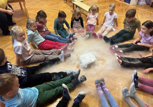 Dzieci siedzą na podłodze z nogami wyciągniętymi, na podłodze leży miska z wodą i suchym lodem, a para unosi się