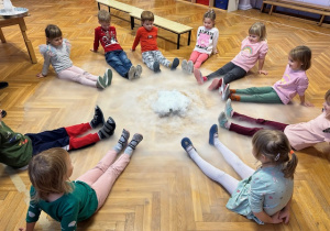 Dzieci siedzą na podłodze z nogami wyciągniętymi a w środku leży miska z wodą i suchym lodem