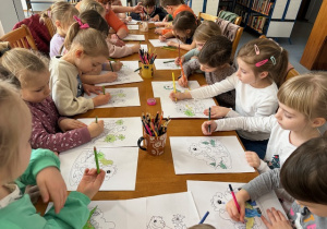 Dzieci siedzą przy stole i kolorują obrazki przedstawiające Dinozaury