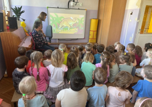 Dzieci siedzą na ławeczkach i oglądają Film edukacyjny o Dinozaurach