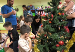 Rodzice i dzieci dekorują choinkę ozdobami- bombkami i łańcuchami