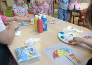 Dzieci siedzą przy stole i malują farbami paluszkiem kropki na parasolce