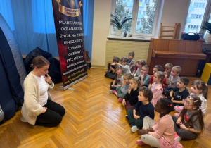 Dzieci siedzą na podłodze i słuchają ciekawych informacji o Kosmosie