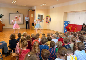 Dzieci siedza na ławeczkach i ogladają taniec baletnic.