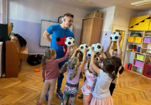 Dzieci stoją z panem Kangurem w kole z piłkami uniesionymi wysoko